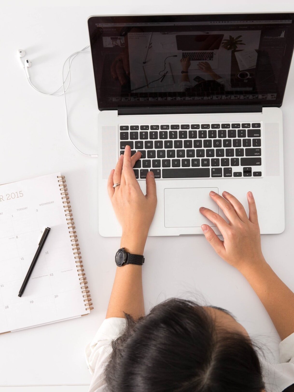 Top view of a person using a laptop on a white desk. A spiral-bound 2015 calendar lies open on the left, and the person's hands are on the keyboard. A watch is visible on their left wrist, and a white charging cable is connected to the laptop.