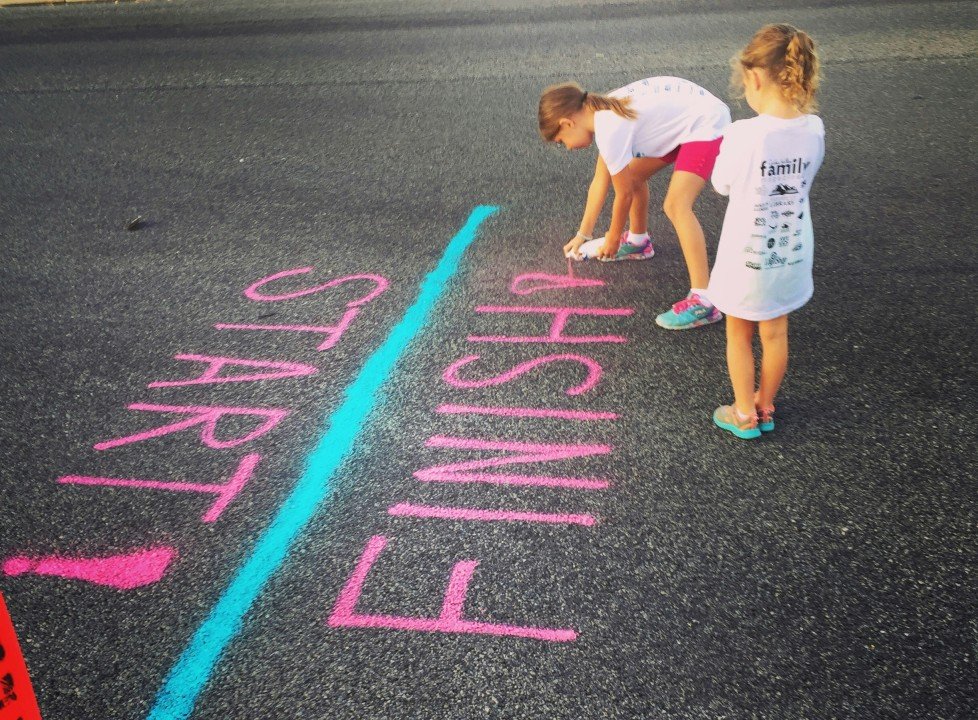 Two young girls stand on a road near brightly painted "START" and "FINISH" lines in pink and blue. One girl kneels, touching the lines, while the other stands nearby. They both wear white shirts and shorts.