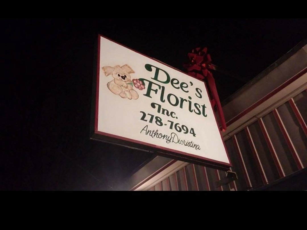 A sign for Dee's Florist Inc., featuring a drawing of a dog holding a flower, with a red ribbon. The text includes a phone number and the name "Anthony D'Ostina" in smaller letters. The sign is illuminated at night.